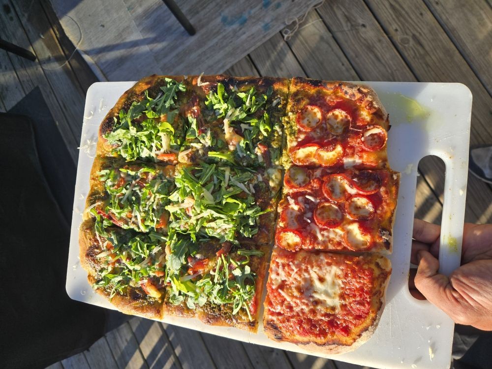A rustic pan pizza with square cuts topped with arugula, pepperoni and fresh cheese on a wooden board outdoors in warm golden-hour light